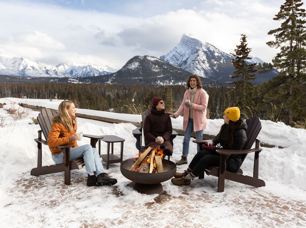 Four friends sit around a campfire in Banff with Rundle Mountain in the background.